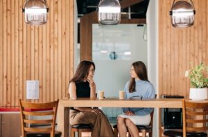 Two corporate women sitting together at a long table drinking iced coffee