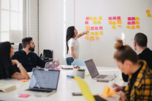 Woman pointing at post it notes on wall, colleagues seated at table in front