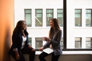 Two women speaking with one another while sitting on a windowsill in an office.